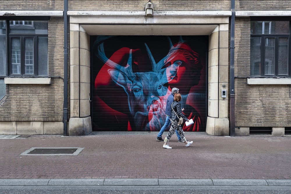 Twee mensen wandelen door Hasselt langs een moderne muurschildering van een hert en een man.